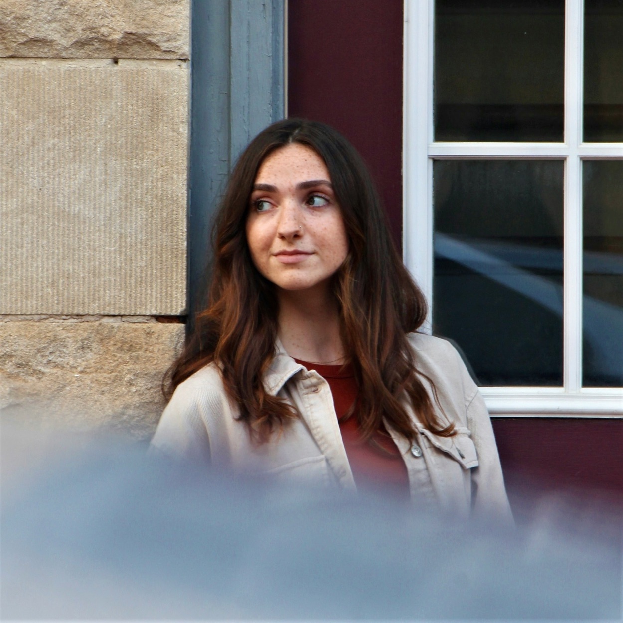 A woman in a tan jacket standing against a cranberry wall, glancing to the side